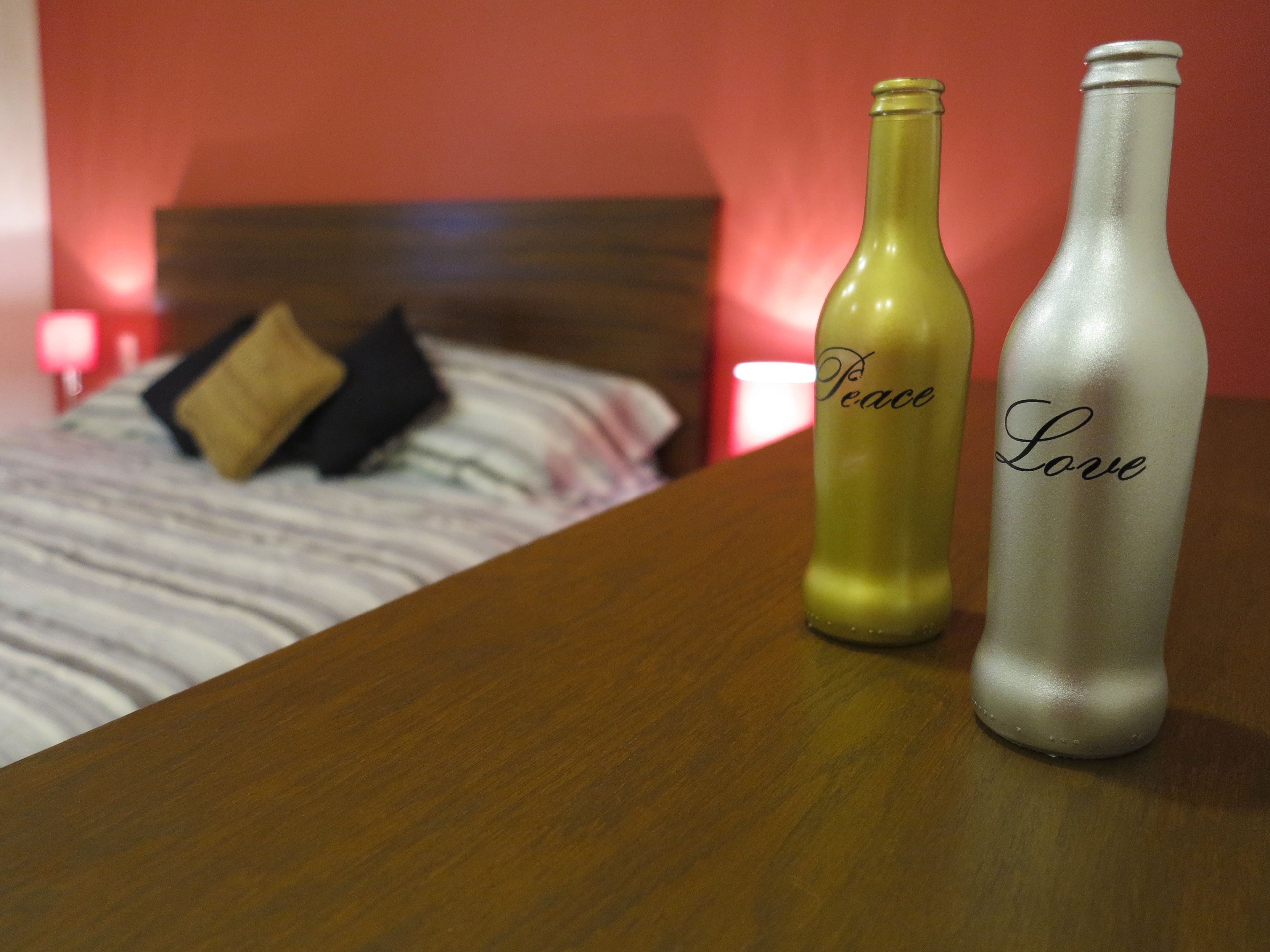 Two bottles labeled "Peace" and "Love" on a wooden table in a room with a bed and pillows in the background.