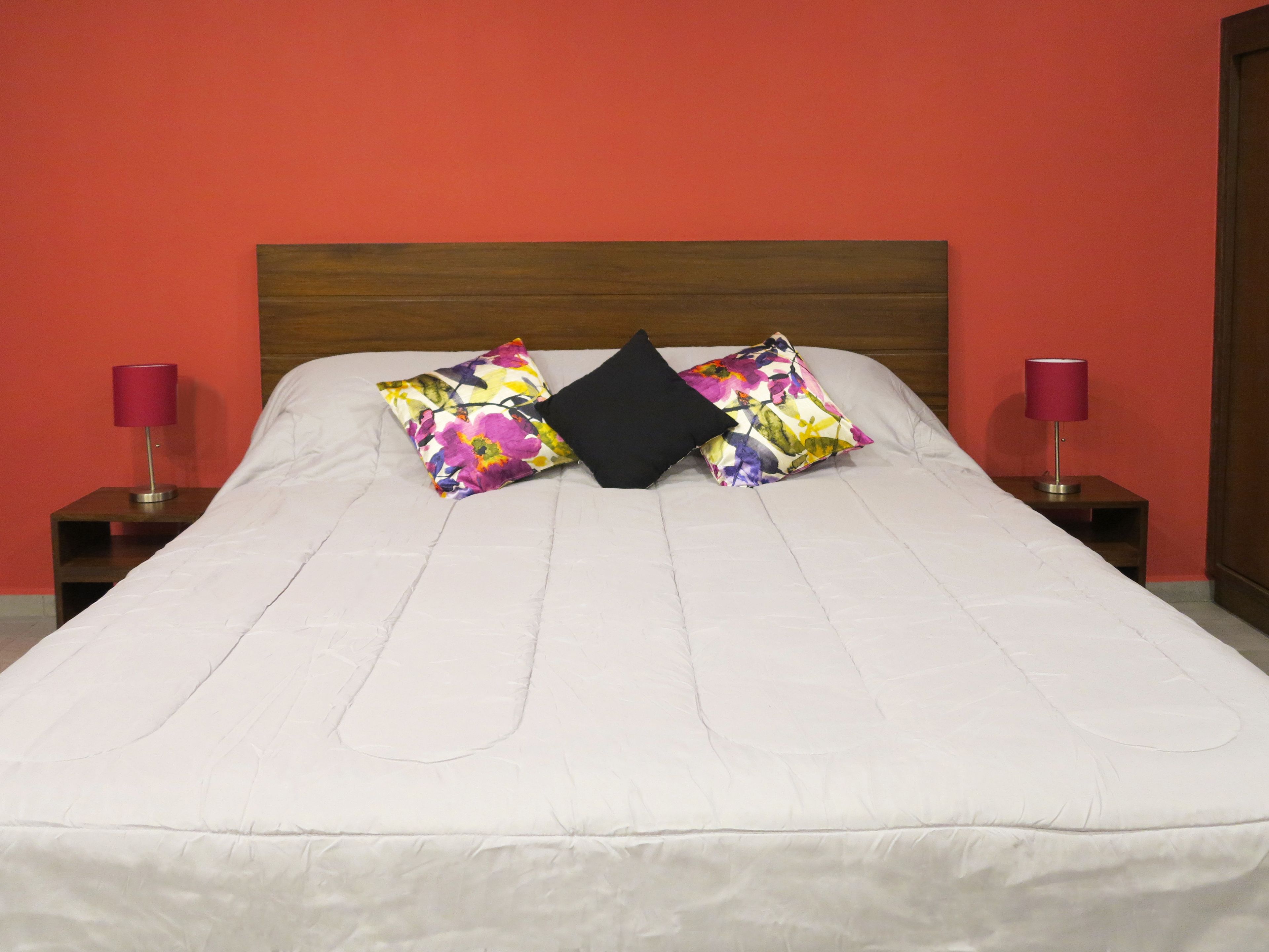 Una cama con edredón blanco, dos almohadas florales y una almohada negra. Hay dos mesitas de noche con lámparas.