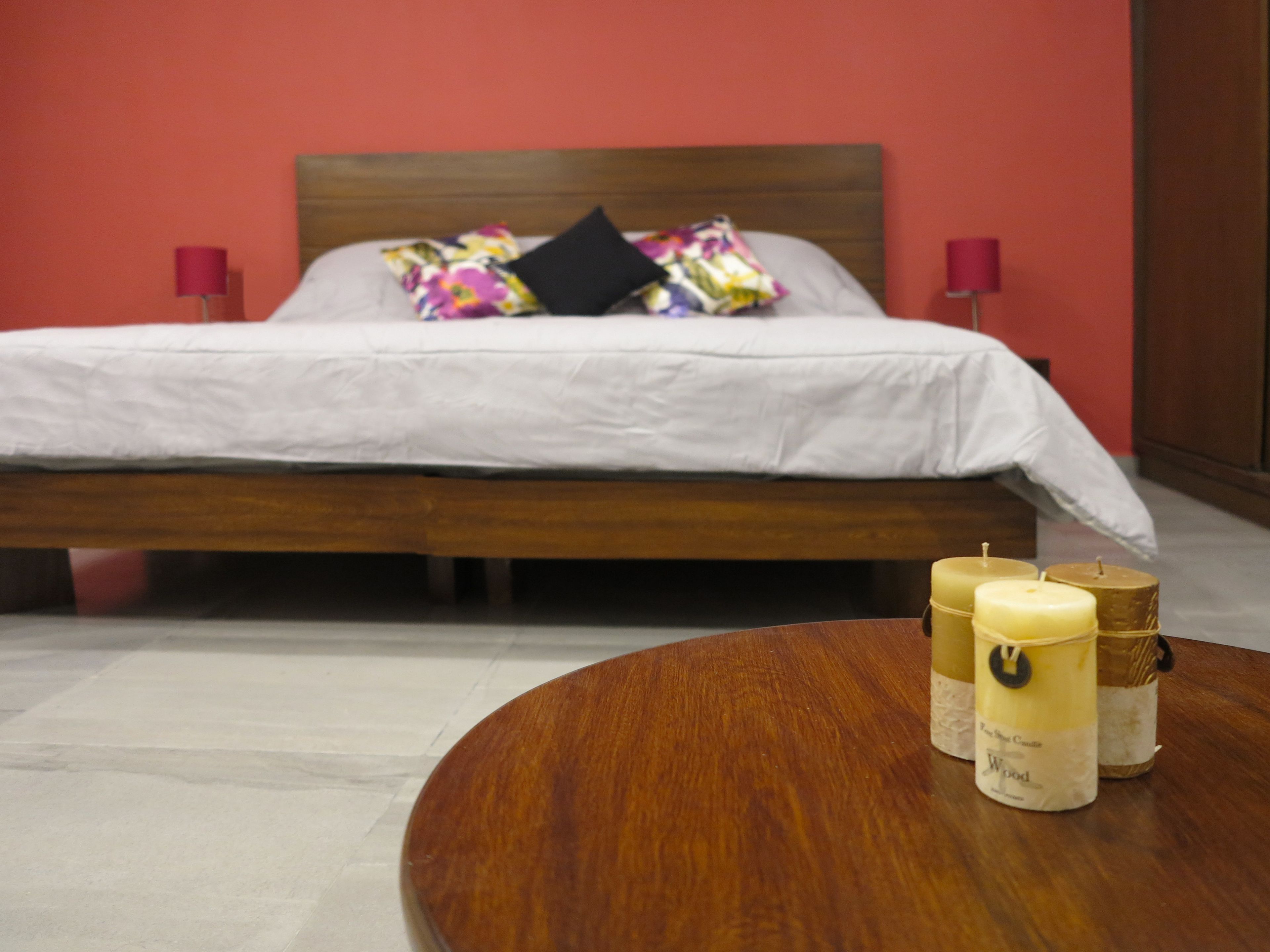 Una cama bien hecha con edredón blanco y almohadas coloridas, una mesa redonda de madera con velas, y una pared roja al fondo.