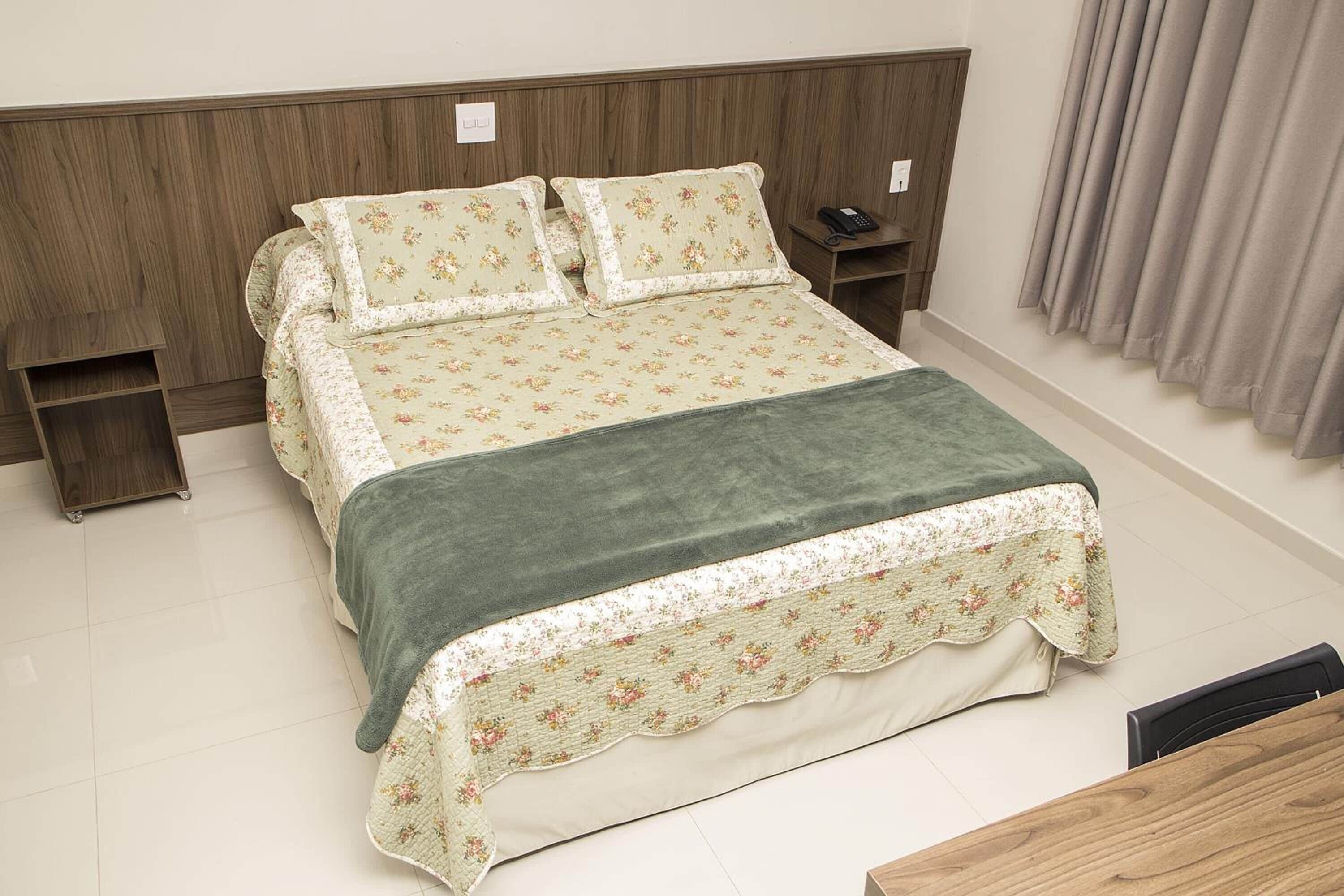 A neatly made bed with floral bedding, a wooden headboard, a bedside table with a phone, and a curtain on the window.