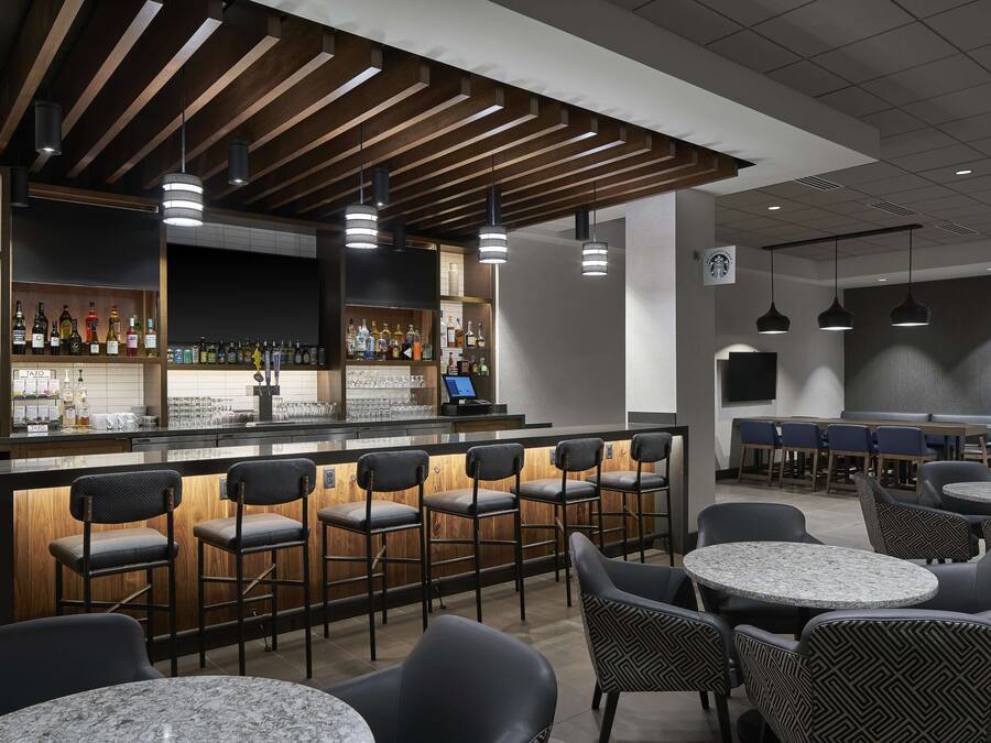 Uma área de bar com balcões de madeira, banquetas e iluminação pendente. Há mesas redondas e cadeiras na área de estar.