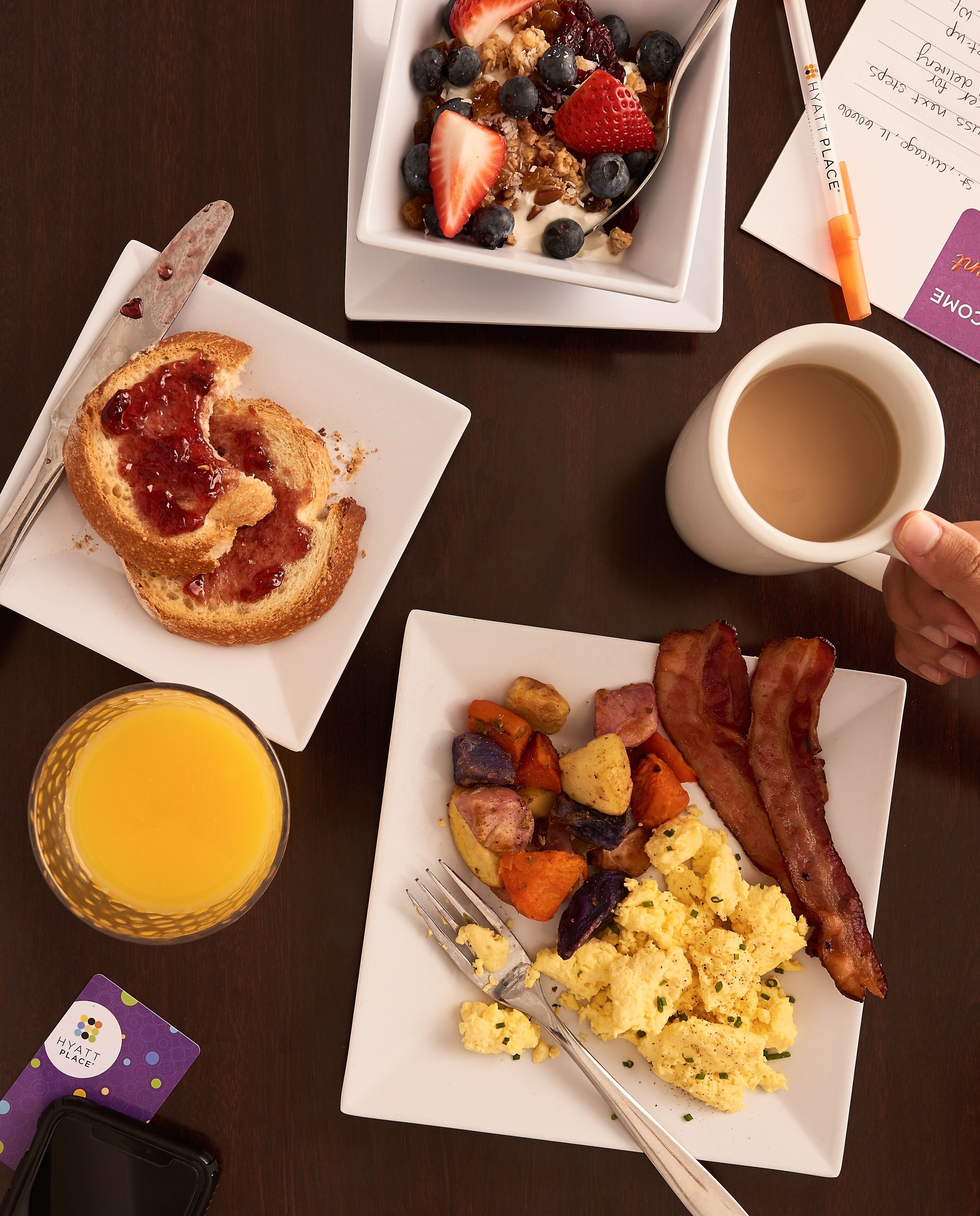 Une table de petit-déjeuner avec des toasts, des œufs brouillés, du bacon, des légumes rôtis et un verre de jus d’orange.