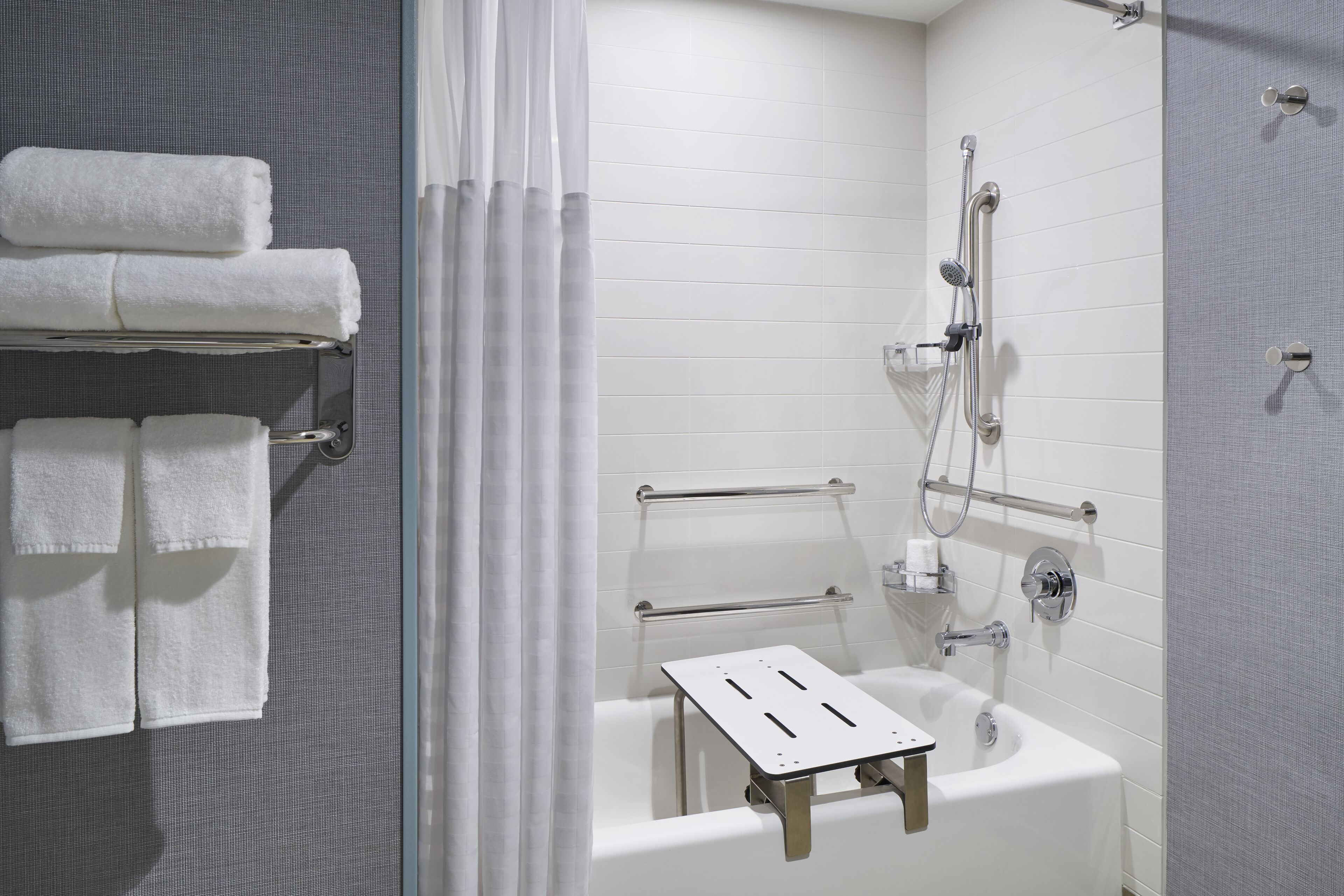A modern bathroom with a bathtub, shower, and handrails. There are towels on a rack and a shower curtain.