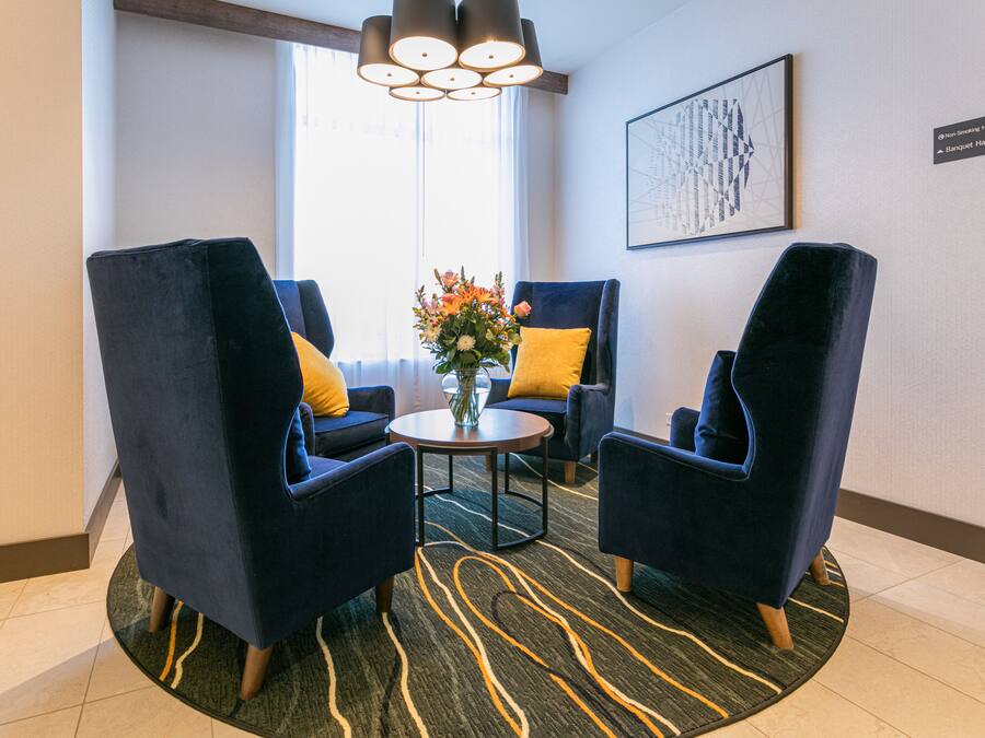 A hotel lobby with two dark blue armchairs, a small round table, and a vase with flowers. A chandelier hangs from the ceiling, and a framed abstract artwork is on the wall.