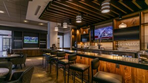 A bar area with wooden bar counter, multiple stools, and a television screen displaying a sports game. - Hyatt Place Wilmington Riverfront (Wilmington)
