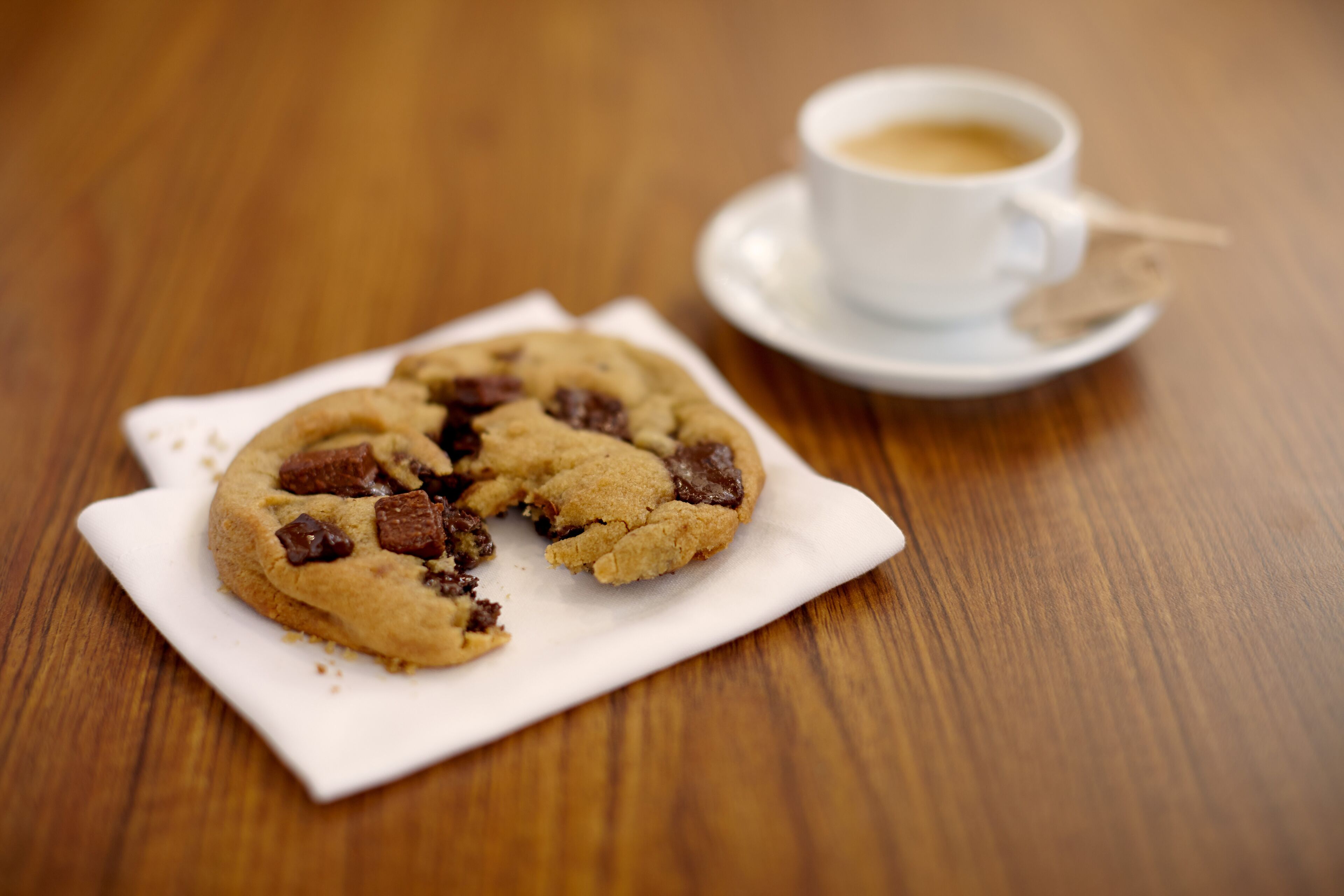 Un biscuit aux morceaux de chocolat, posé sur une assiette blanche, à côté d’une tasse de café sur un sous-tasse.