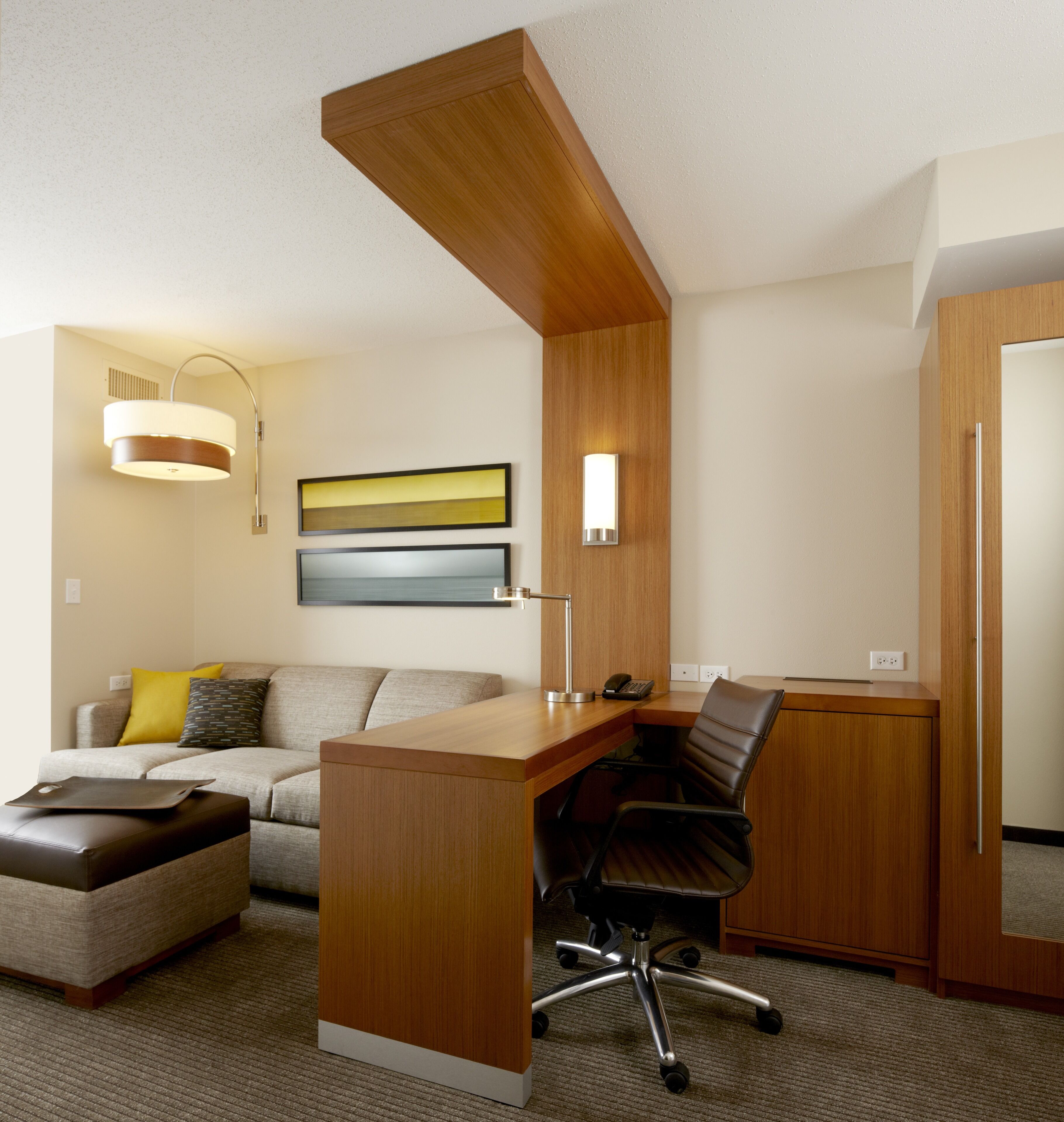 Une chambre d’hôtel moderne avec un bureau en bois, un fauteuil de bureau noir, un canapé avec des coussins, une lampe fixée au mur et un tableau.