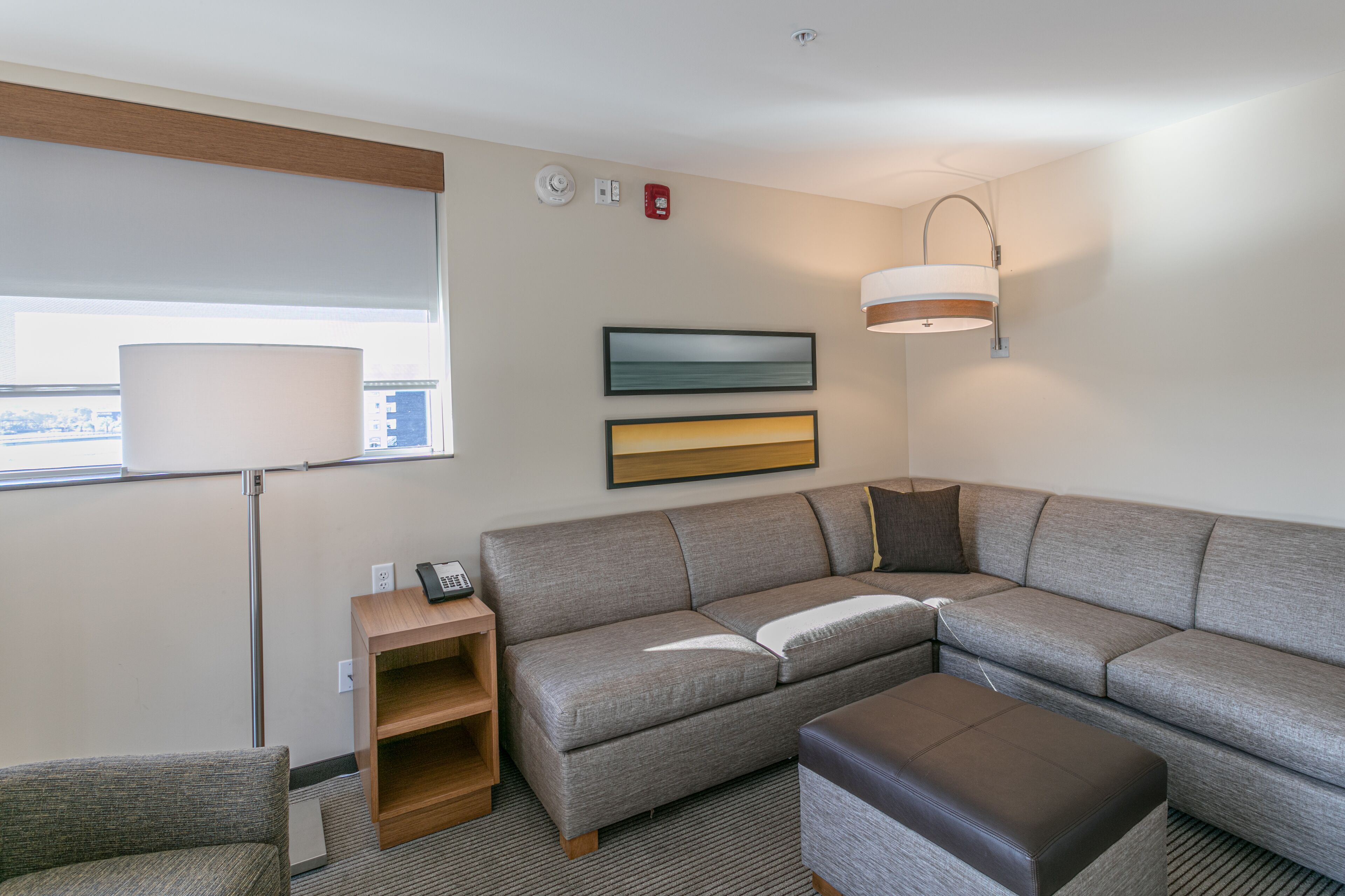A modern hotel room with a sectional sofa, a small wooden side table, a lamp, and a large window with a view.