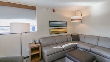 A modern hotel room with a sectional sofa, a small wooden side table, a lamp, and a large window with a view.