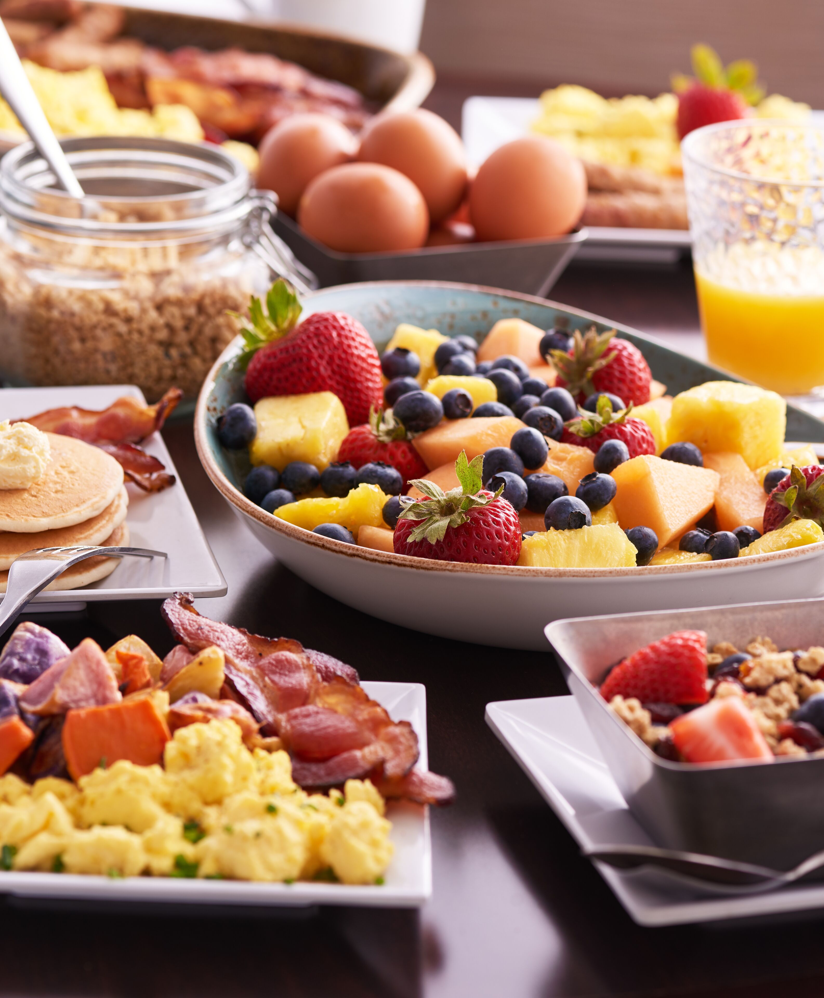A breakfast spread with various dishes including scrambled eggs, bacon, pancakes, and a fruit bowl with strawberries, blueberries, and pineapple.