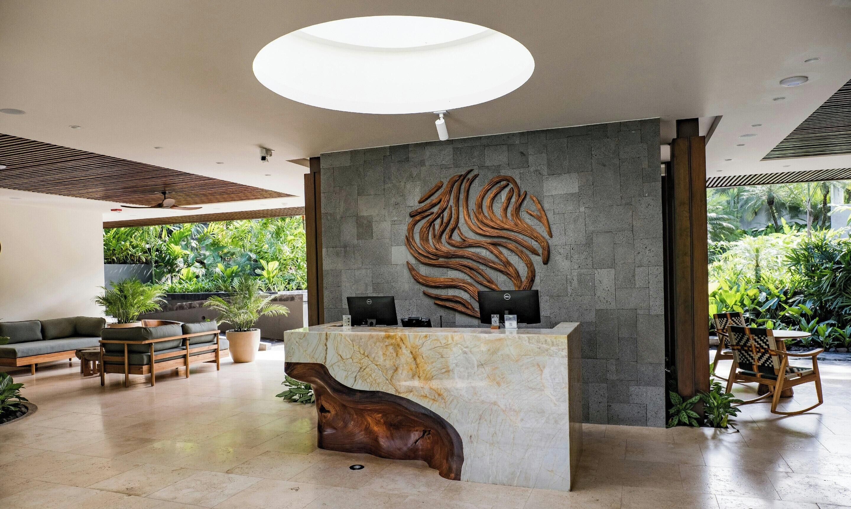 A reception desk with a unique wood carving, a modern ceiling light, and a view of lush greenery outside.
