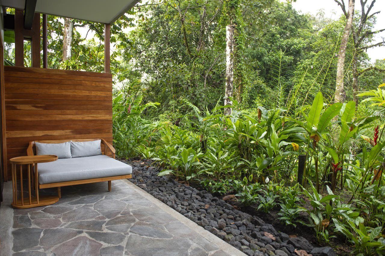 A wooden bench with cushions and a small table, surrounded by a lush garden with various plants and a stone pathway.
