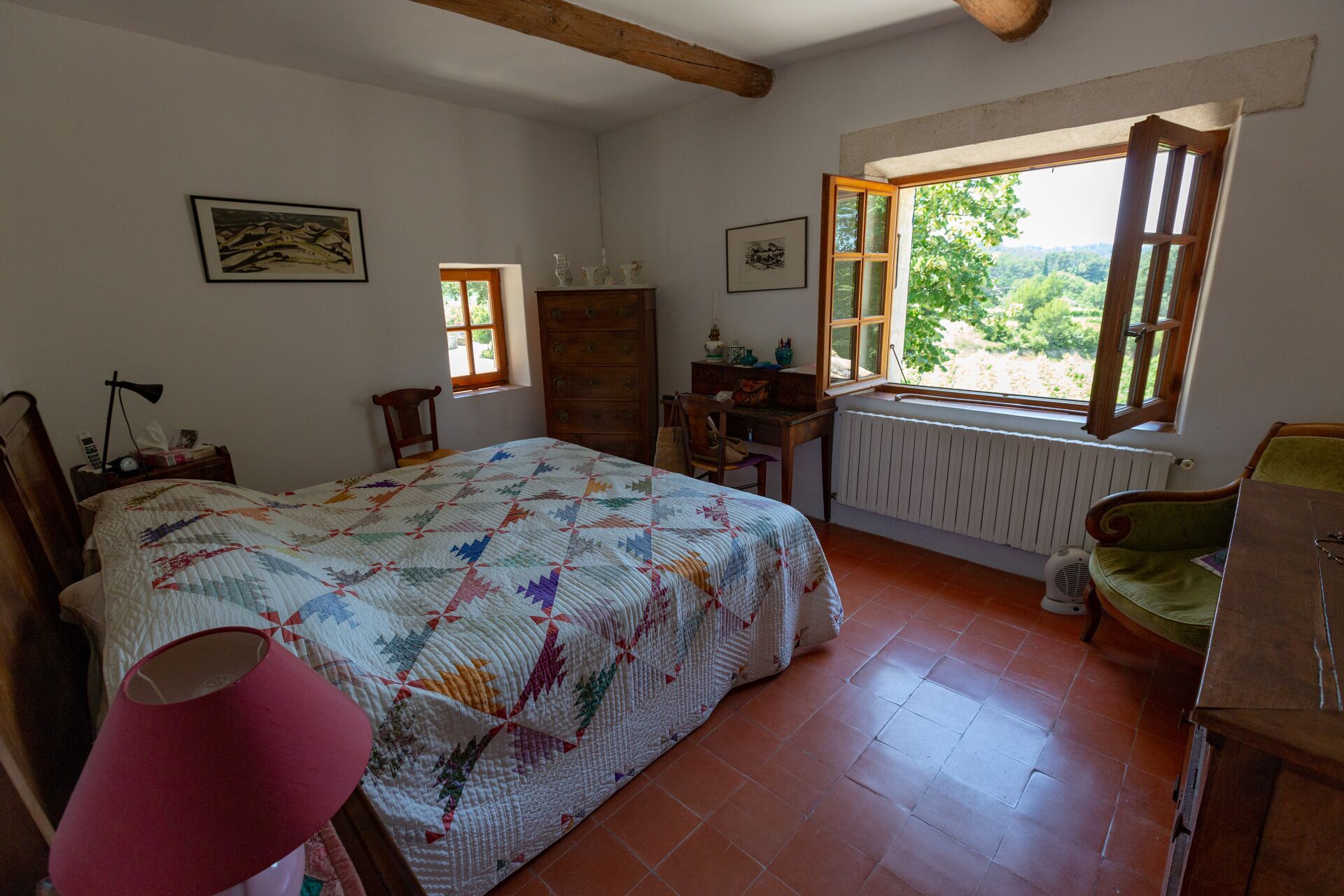 A bedroom with a large bed, a desk, a chair, a radiator, and a window with a view of greenery.