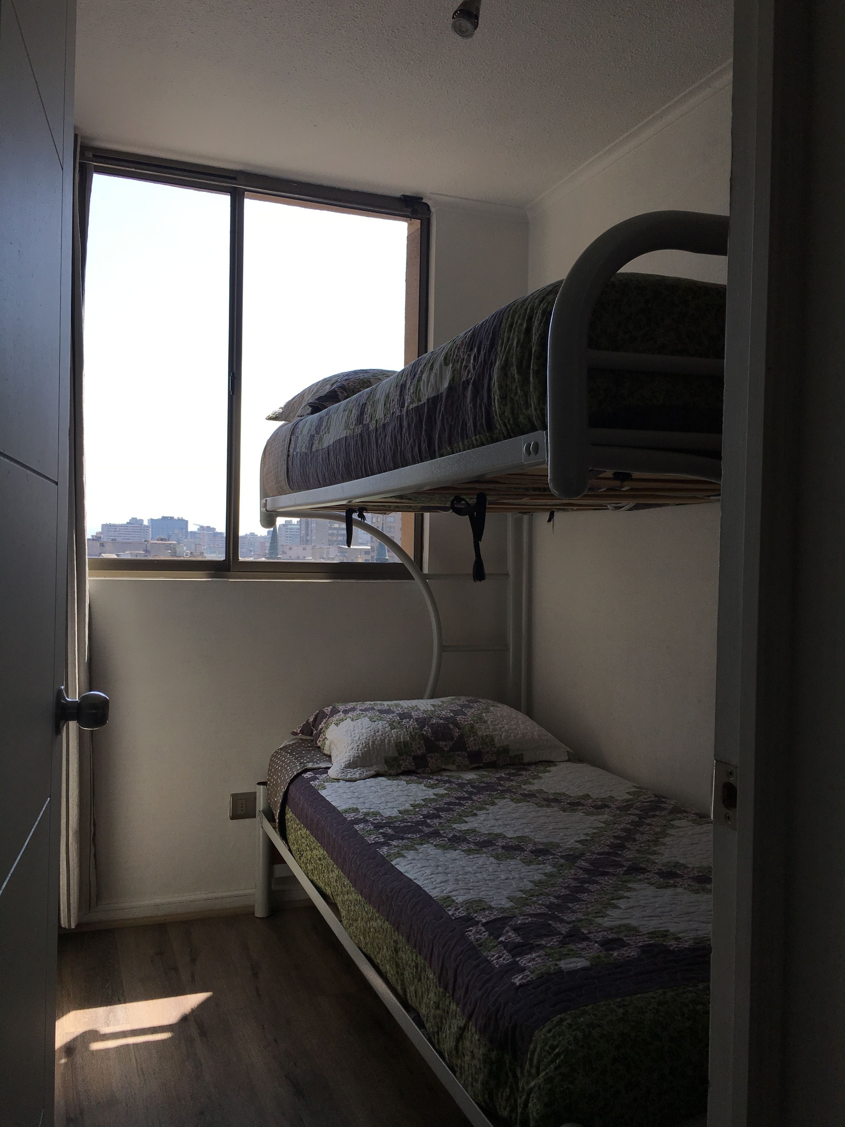 Ein kleines Zimmer mit Etagenbett, einem Fenster und Stadtblick sowie einer Tür.