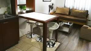 A compact living space with a kitchenette, a dining area with two stools, a sofa with cushions, a fan, and a small table. - Amistar Apartments (Santiago)