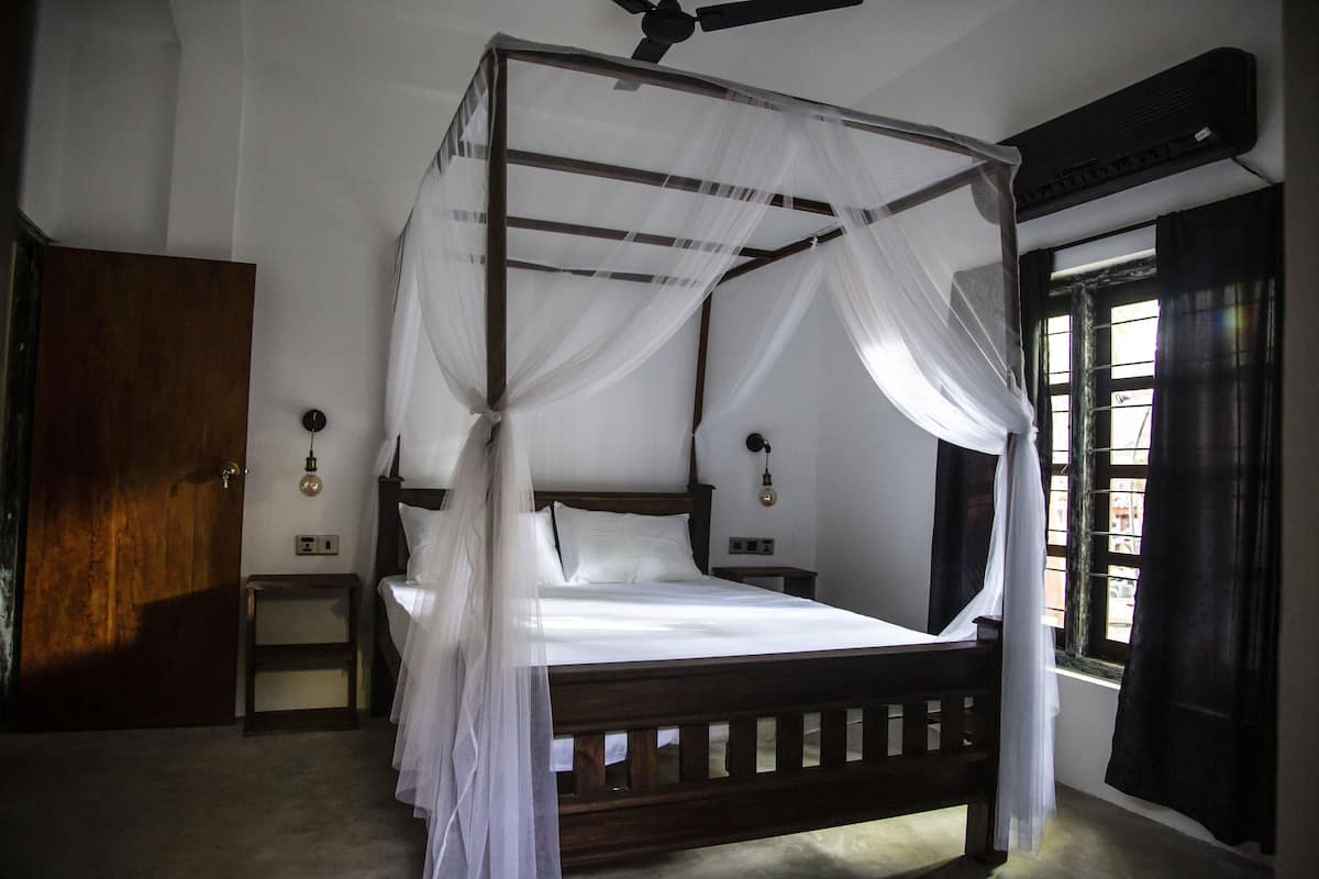Uma cama com dossel e cortinas, roupa de cama branca, cabeceira de madeira e mesa de cabeceira com abajur.
