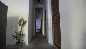 A narrow hallway with a potted plant, a wooden door, and a view into a room with a bed.