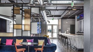 A modern lounge area with a blue sofa, two round tables, and a bar with stools. - Aloft by Marriott Austin Round Rock (Round Rock)