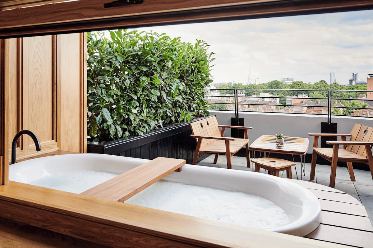 A modern bathroom with a wooden bathtub, a black planter with green foliage, and wooden chairs and tables on a balcony with a city view.