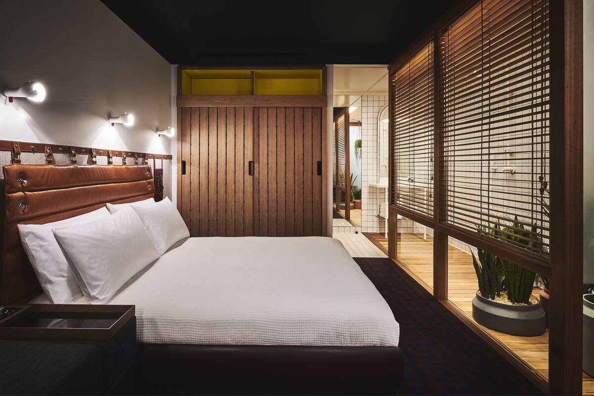 A modern bedroom with a wooden headboard, a bed with white linens, a nightstand, and a view through a wooden partition to a bathroom.