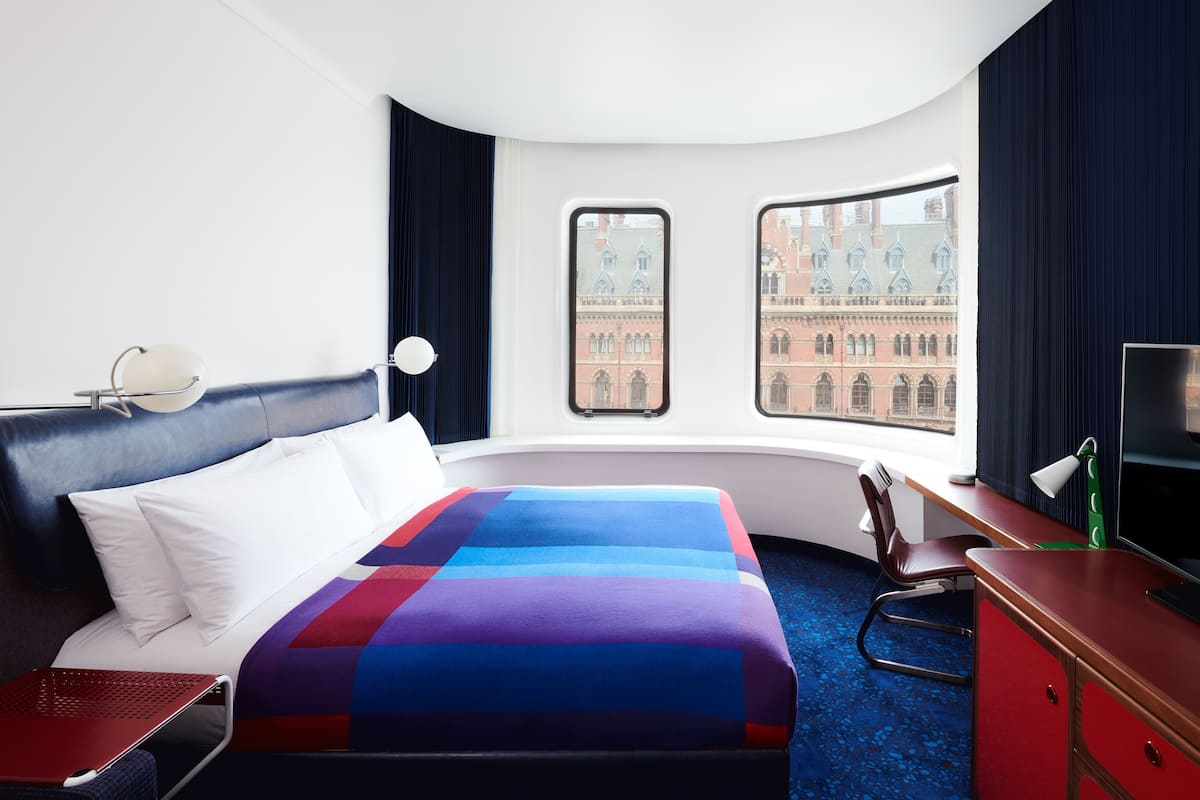 A hotel room with a bed, a desk, a chair, a TV, and a window with a view of a building.