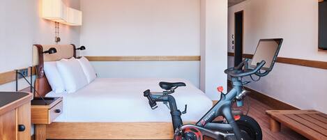 A hotel room with a bed, a desk, a chair, a standing desk, a laptop, and a stationary exercise bike.