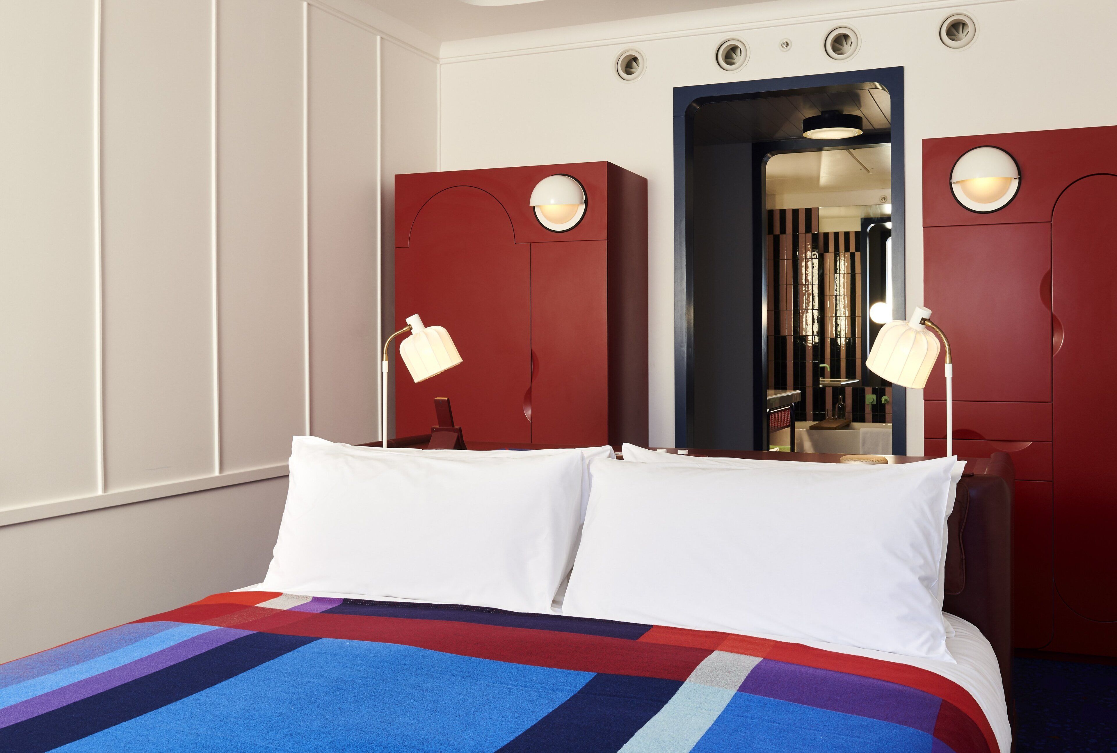 Una habitación de hotel con una cama grande, dos lámparas de noche, un armario rojo y un espejo.