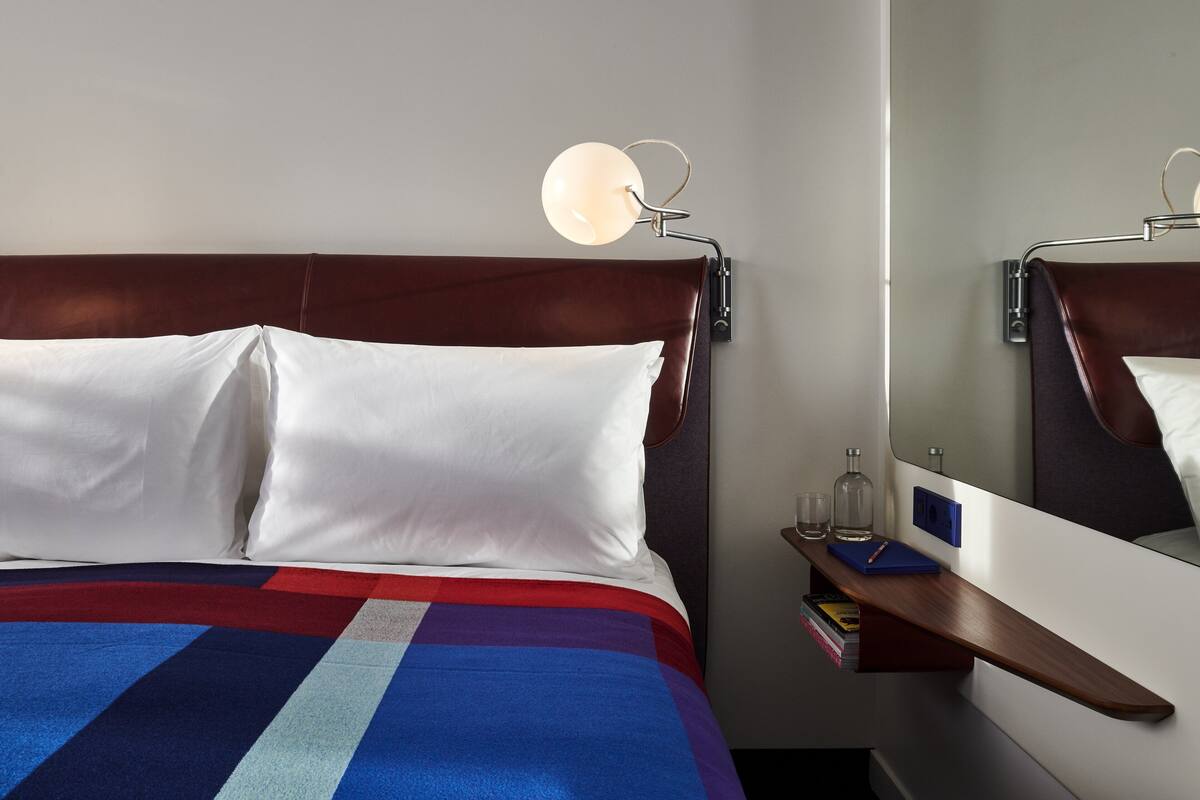 A neatly made bed with white pillows and a multicolored blanket, a bedside table with a lamp, and a mirror on the wall.