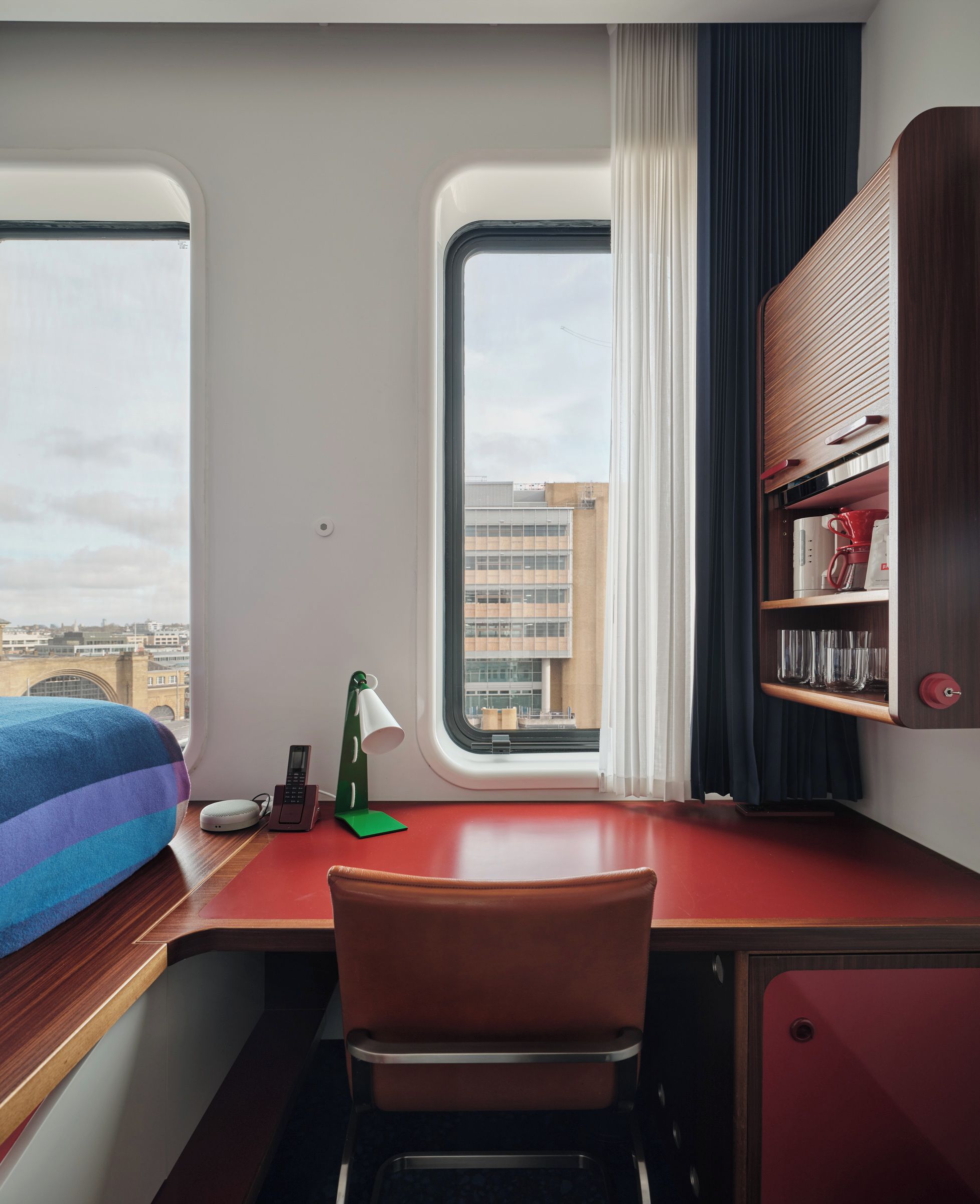 A compact room with a bed, a desk, a chair, a window with a view of buildings, and a shelf with items.