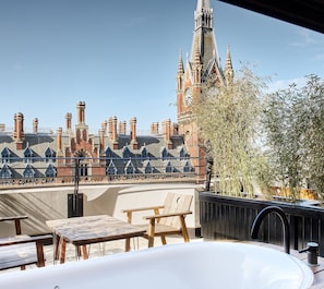 A rooftop terrace with a view of a historic building, including a clock tower. There are wooden chairs and a table, and a large planter with bamboo.
