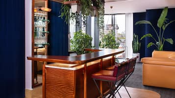 A bar area with a wooden counter and red cushioned stools, a seating area with a yellow sofa, and large windows with plants.