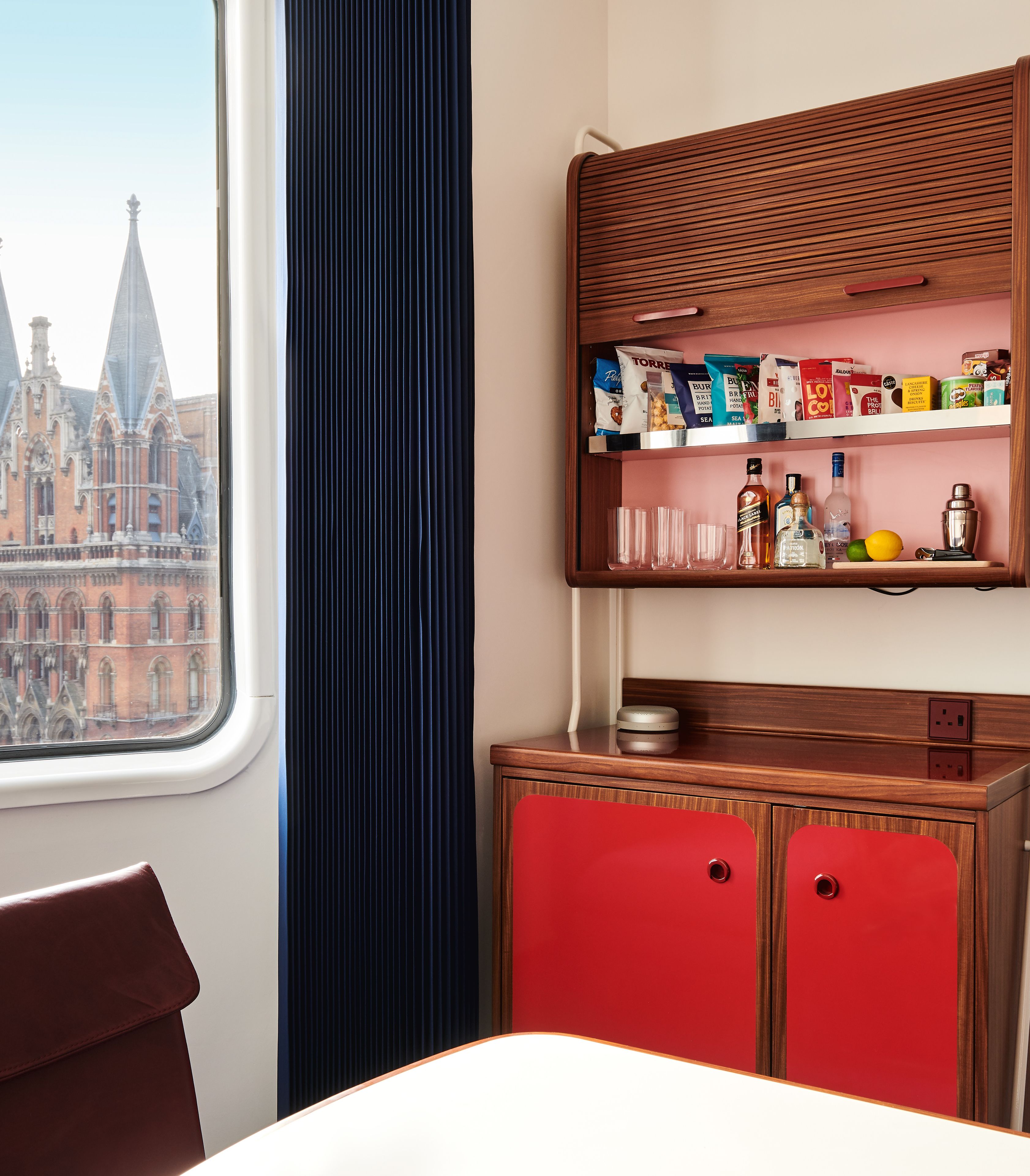 Une chambre d’hôtel avec vue sur un bâtiment historique, un buffet en bois rempli de collations et un fauteuil rouge.