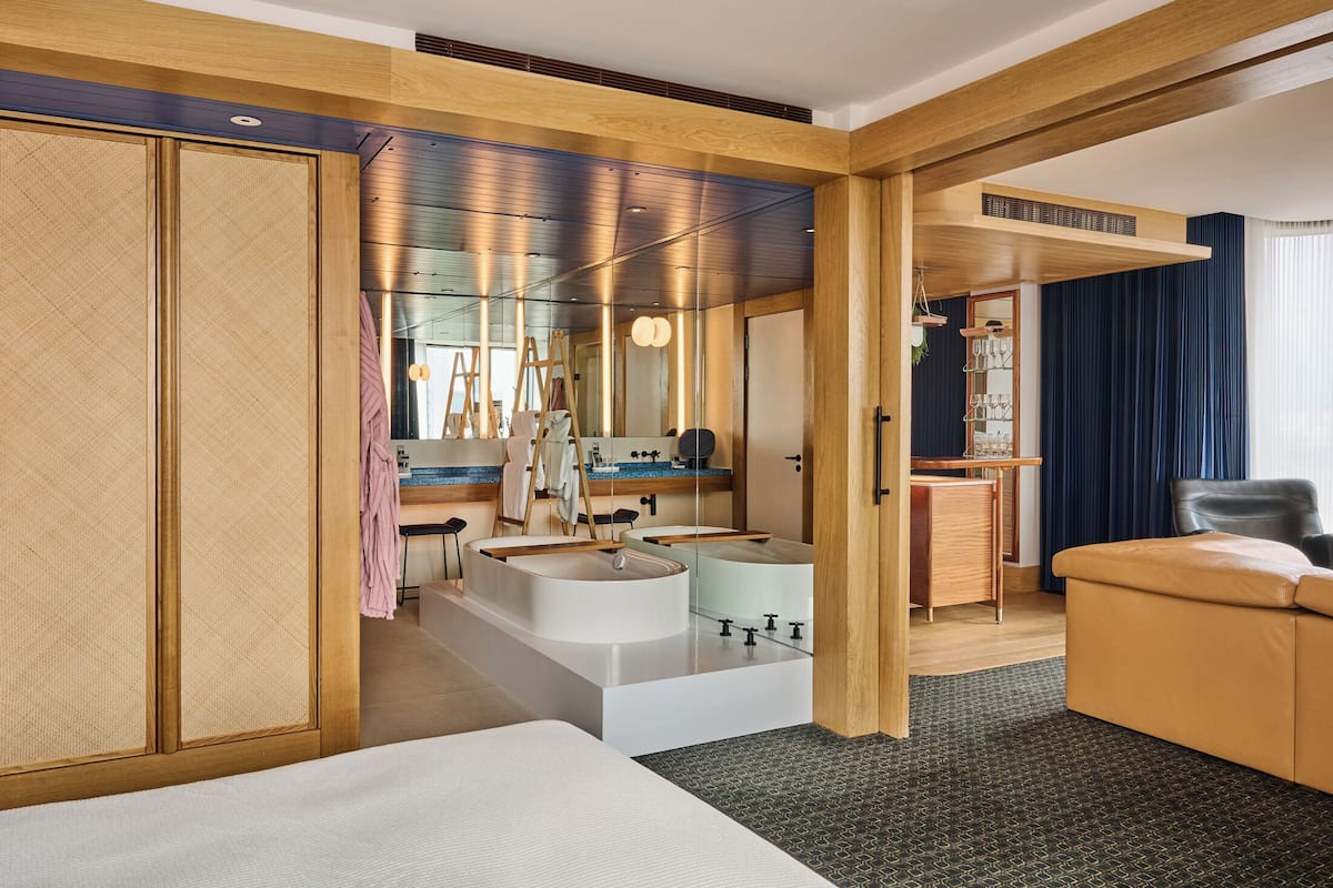 A modern hotel room with a large bed, a sitting area with a sofa, a desk, and a large mirror.