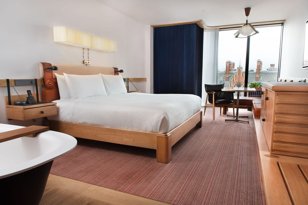 A modern hotel room with a large bed, a desk, a chair, and a view of the city through the window.