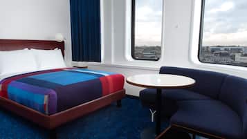 A hotel room with a bed, a seating area, a small table, and large windows offering a city view.
