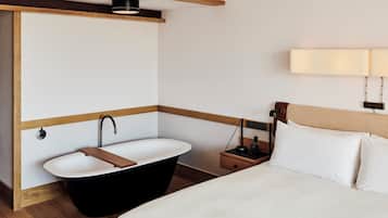 Et moderne hotelværelse med en stor seng, et fristående badekar, et natbord med lampe og en vægmonteret lysarmatur.