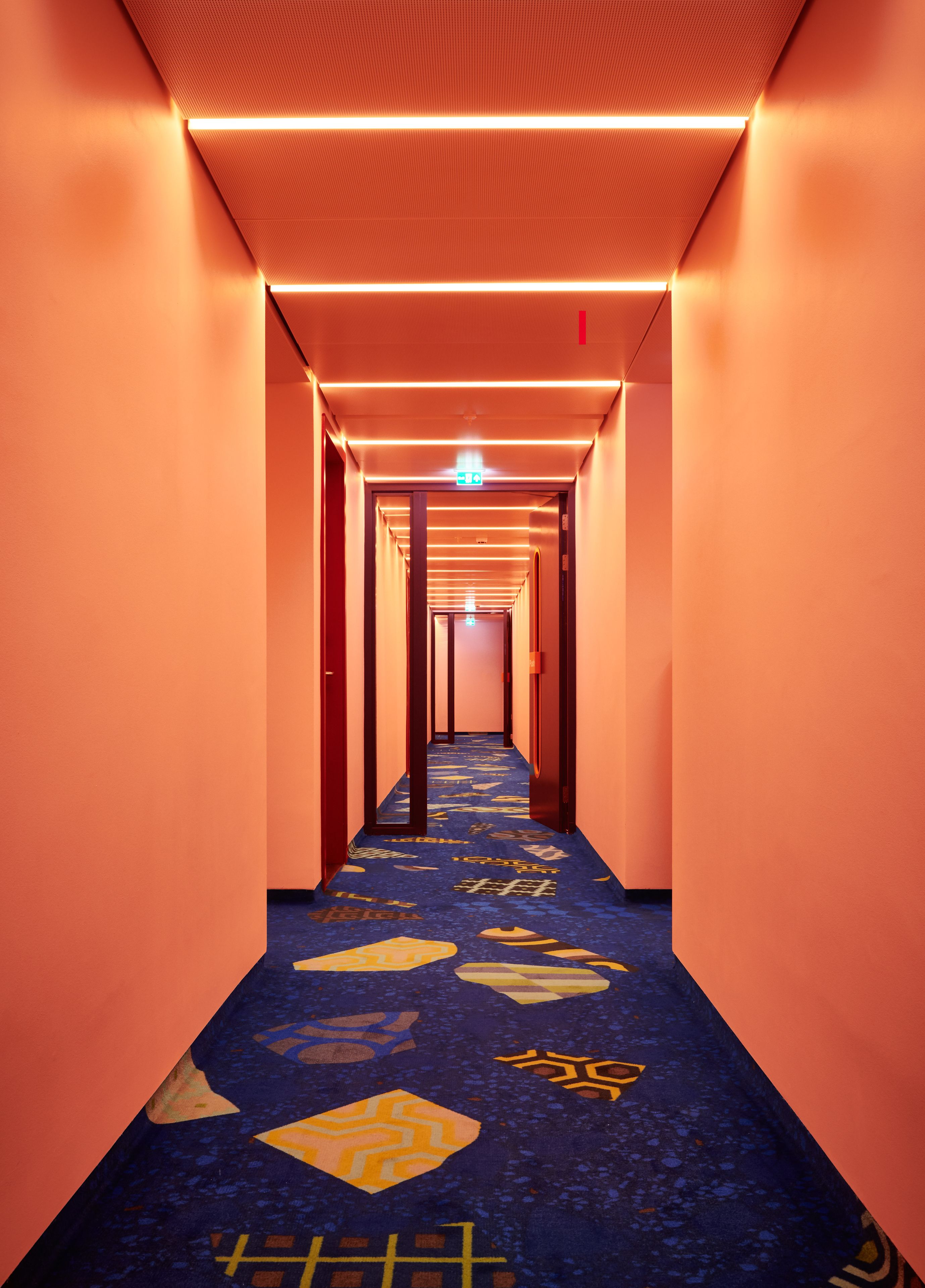 Un corridoio di un hotel con tappeto a motivi blu, pareti arancioni e un soffitto con illuminazione lineare.