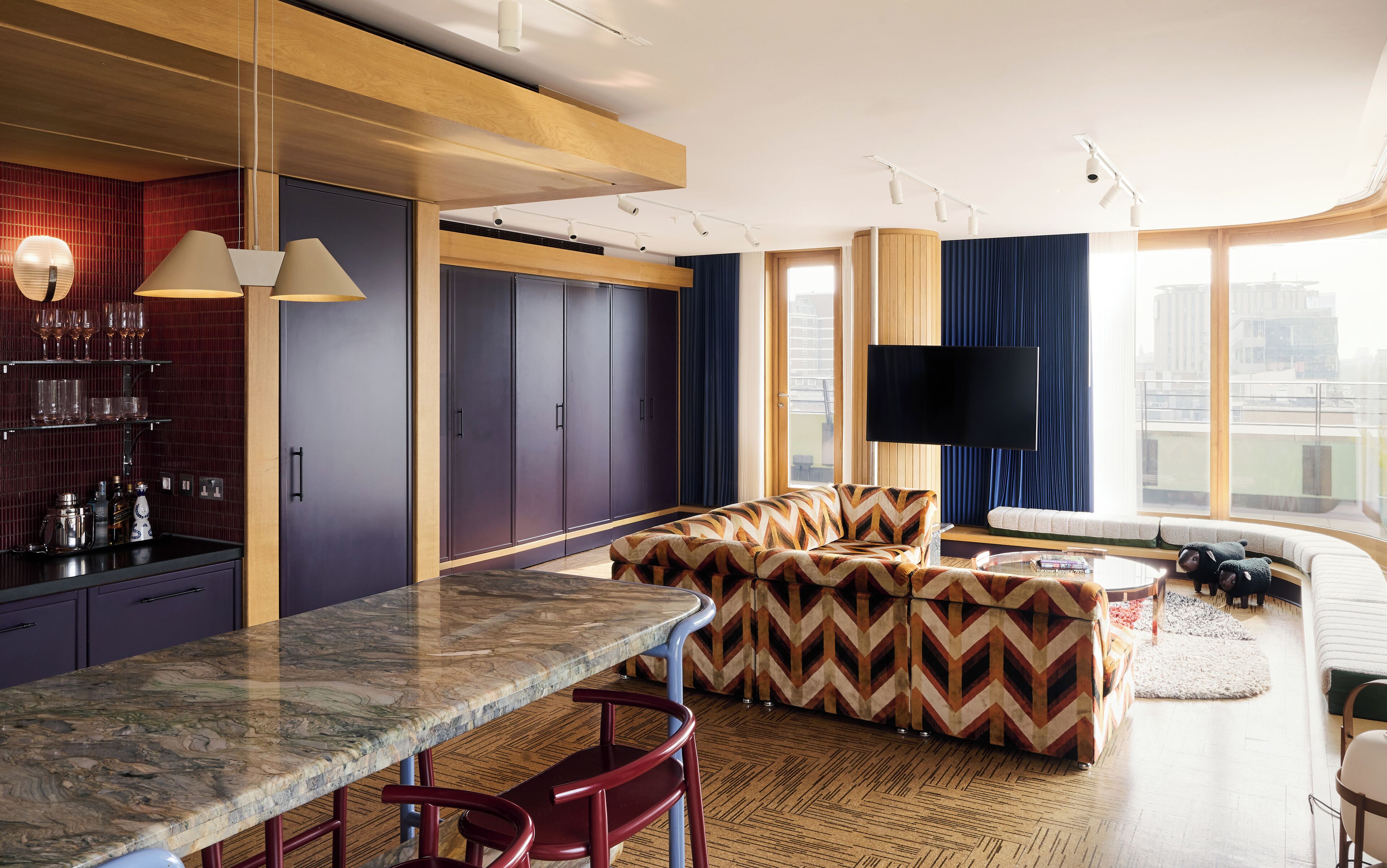 A modern living room with a large TV, a sofa, a coffee table, and a kitchen area with a marble countertop and bar stools.