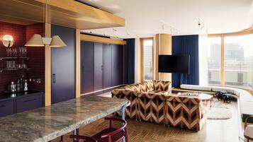 A modern living room with a large TV, a sofa, a coffee table, and a kitchen area with a marble countertop and bar stools.