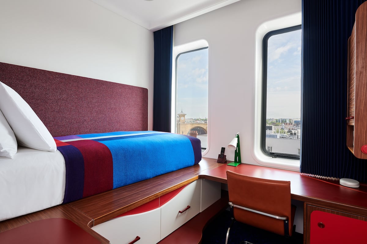 A modern hotel room with a bed, a desk, and a view of a cityscape through a window.