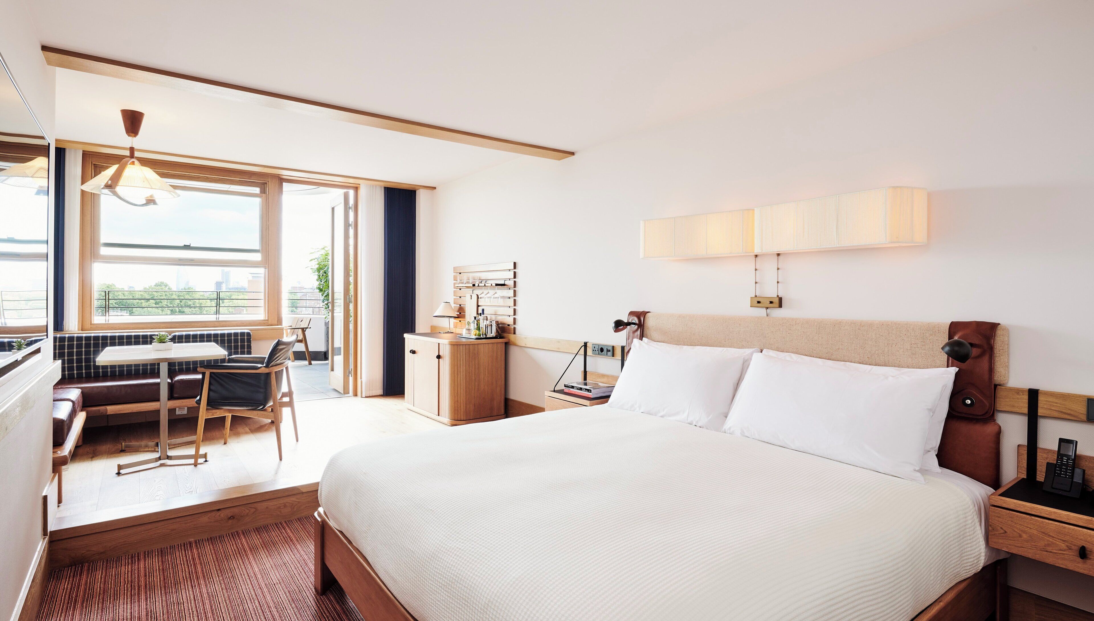 A hotel room with a large bed, a desk with a chair, a small table, a window with a view, and a bedside table with a lamp.