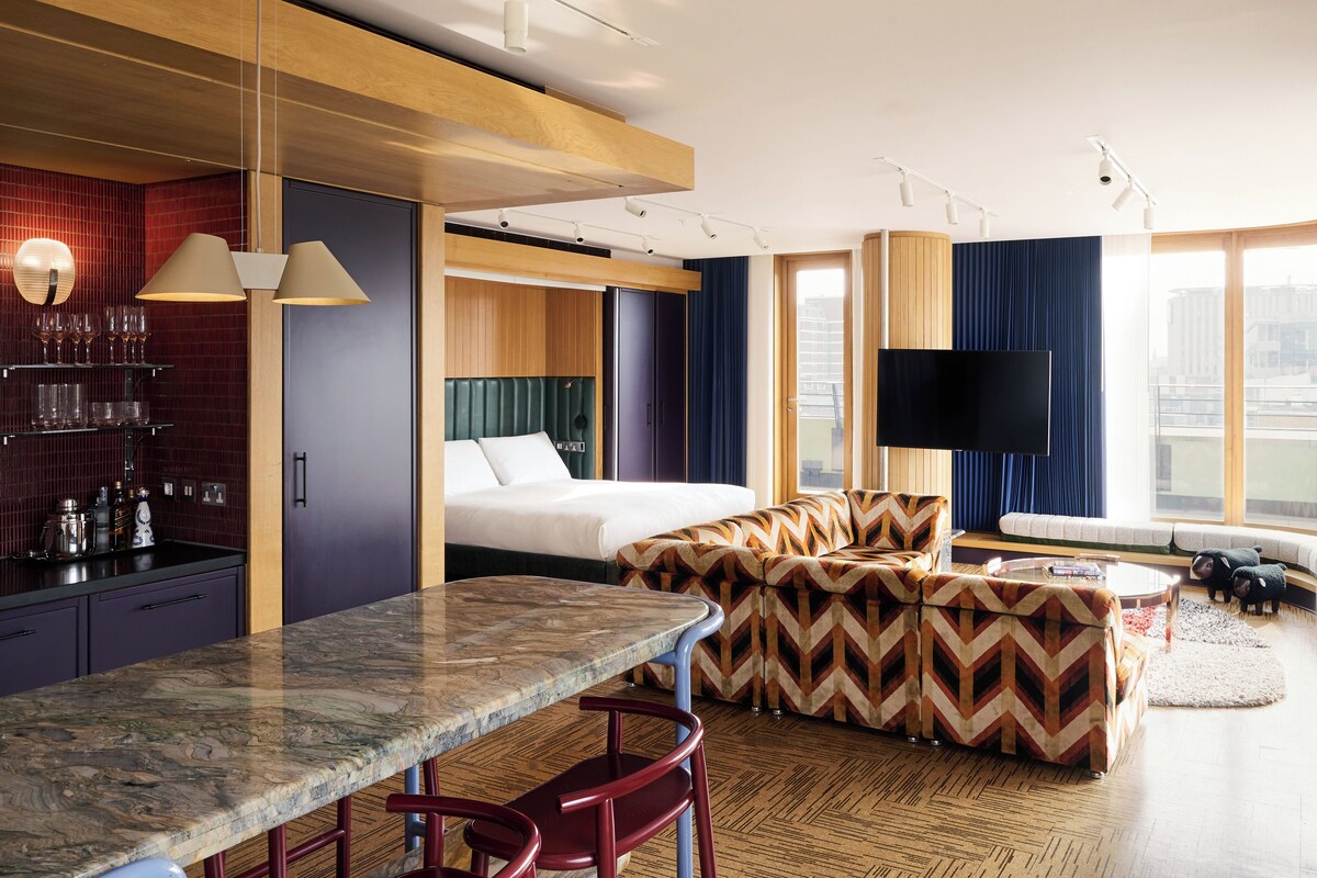 A modern hotel room with a bed, seating area, dining table, and a TV.
