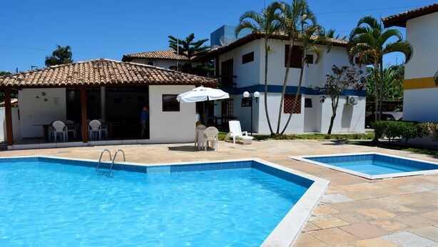 Une piscine avec une terrasse carrelée, un espace de repos couvert avec des chaises, et un bâtiment doté d’un toit carrelé.