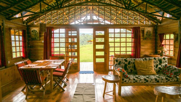 A wooden cabin interior with a dining table, chairs, a sofa, and a view of the outdoors through large windows. - Castle Forest Lodge (Kimunye)