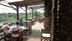 An outdoor wooden deck with tables and chairs, surrounded by lush greenery and a stone wall. - Castle Forest Lodge (Kimunye)