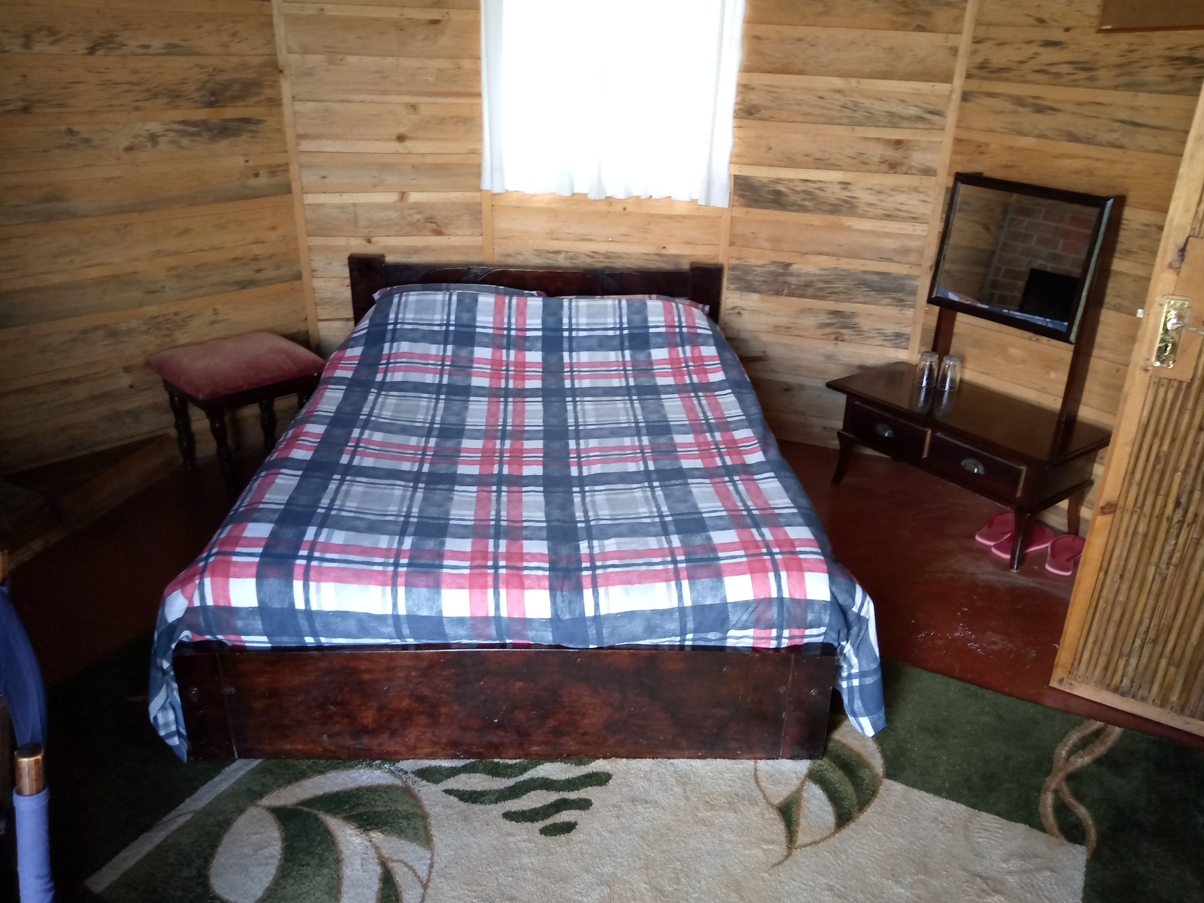 A wooden cabin room with a plaid bedspread, a small wooden bench, a nightstand with a mirror, and a green carpet with a leaf pattern.