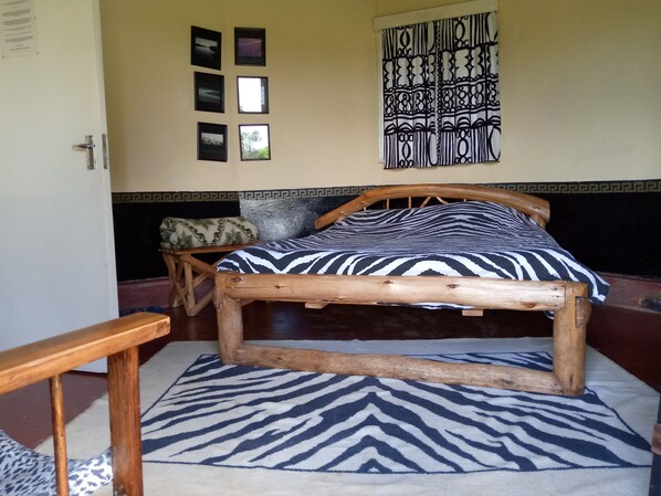 A room with a wooden bed, a patterned rug, a small wooden table, and a wall with framed pictures and a decorative tapestry. - Castle Forest Lodge (Kimunye)