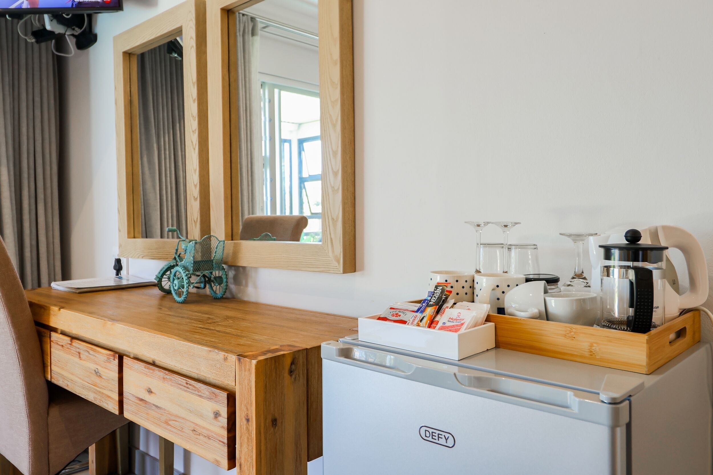 A compact hotel room with a wooden desk, a small refrigerator, and a mirror above it.