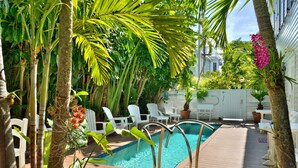 A pool area with a wooden deck, white chairs, and lush greenery including palm trees and flowering plants. - STARS AND STRIPES ~ Magnificent Home Located In The Heart of Old Town! (Key West)