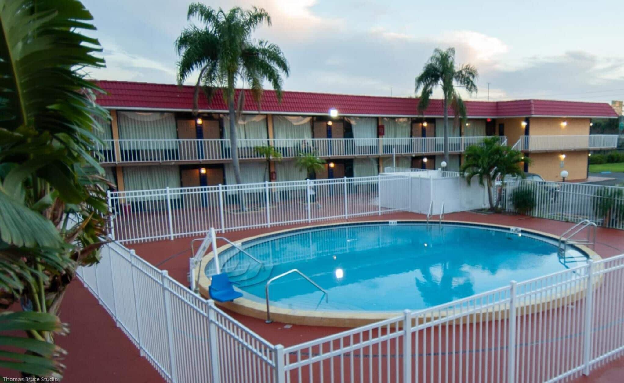 Un hotel con piscina rodeado por una valla blanca, palmeras y un edificio de techo rojo.